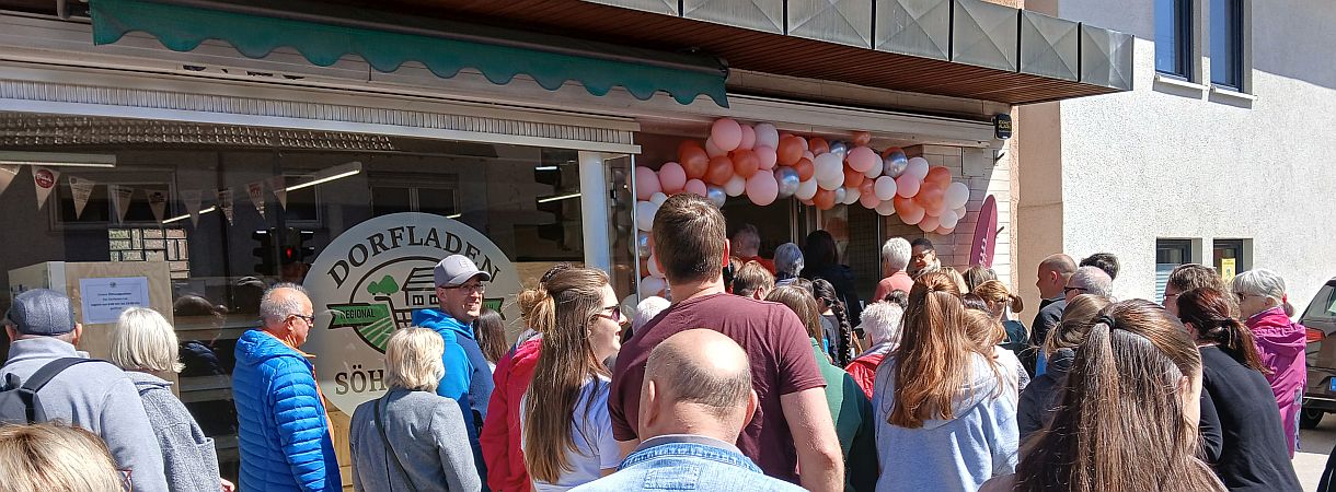 Viele Menschen strömen zum Eingang des Dorfladens, der mit Luftballons geschmückt ist