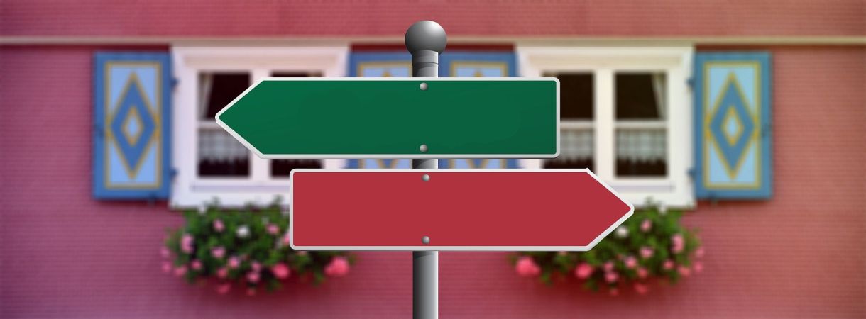 Schild mit einem grünen Pfeil der nach links zeigt und einem roten Pfeil der nach rechts zeigt. Im Hintergrund eine Hausfasade mit Fenstern,  Fensterläden und Blumenkübeln