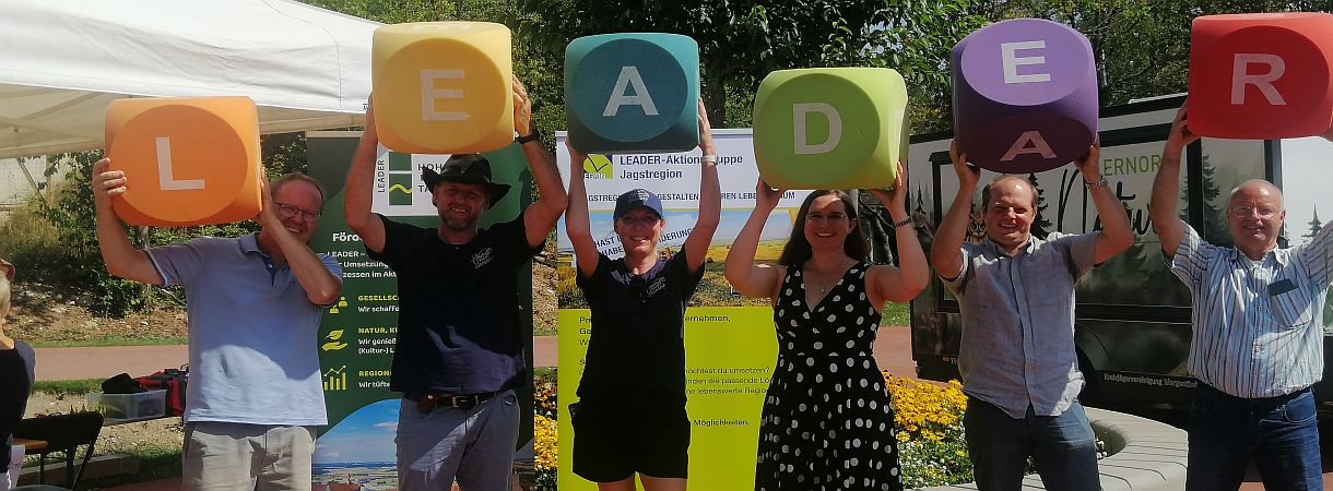 sechs Menschen im Freien, jeder hält einen großen farbigen Würfel mit den Buchstaben L E A D E R 